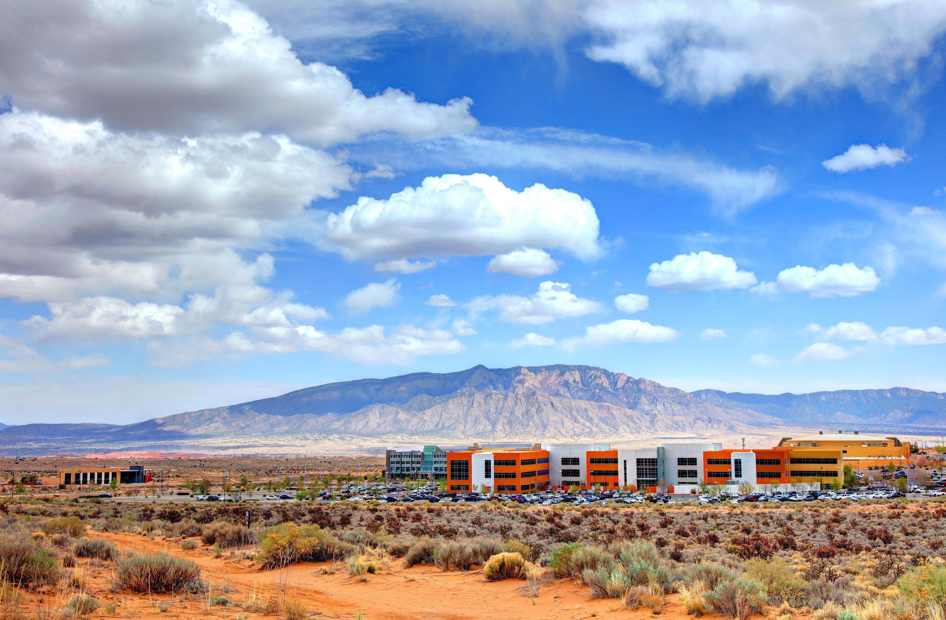 Wide landscape of Rio Rancho’s low-rise office complex set in the high desert, with the Sandia Mountains rising under a bright blue sky dotted with fluffy clouds.