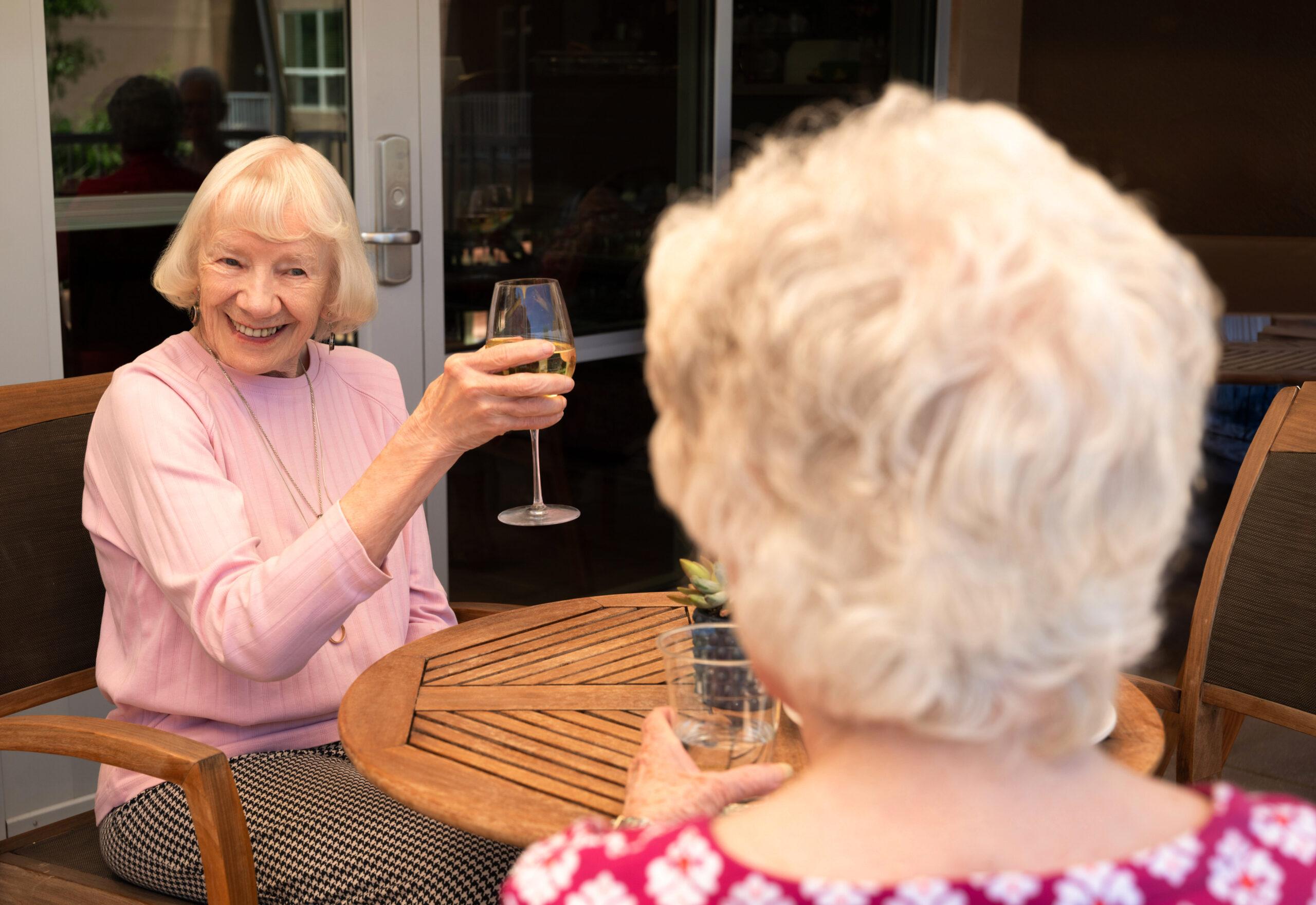 Two older women seated at a round wooden patio table outdoors raising glasses of white wine in a toast, one facing the camera.