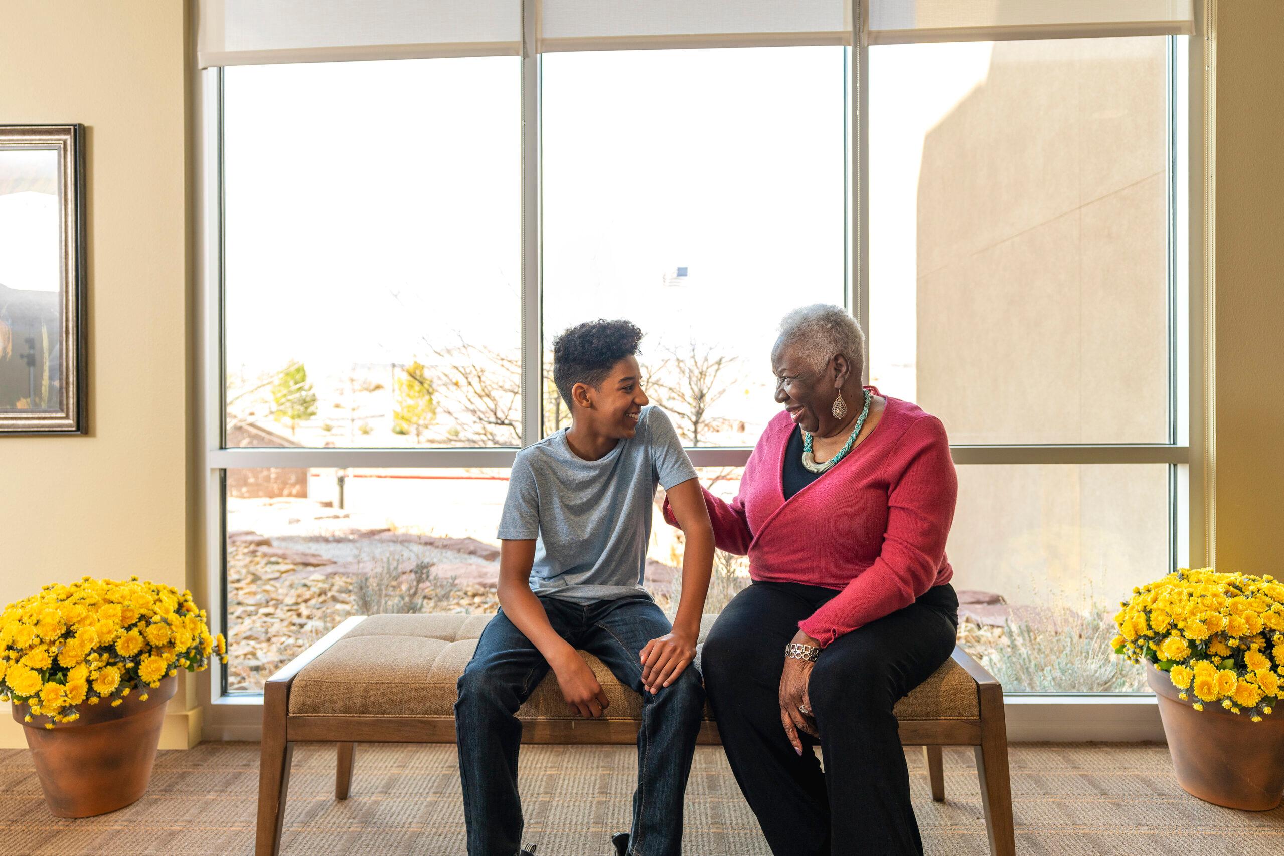 A teenage boy and an elderly woman sitting on a bench in front of a window, smiling and conversing between yellow potted flowers.
