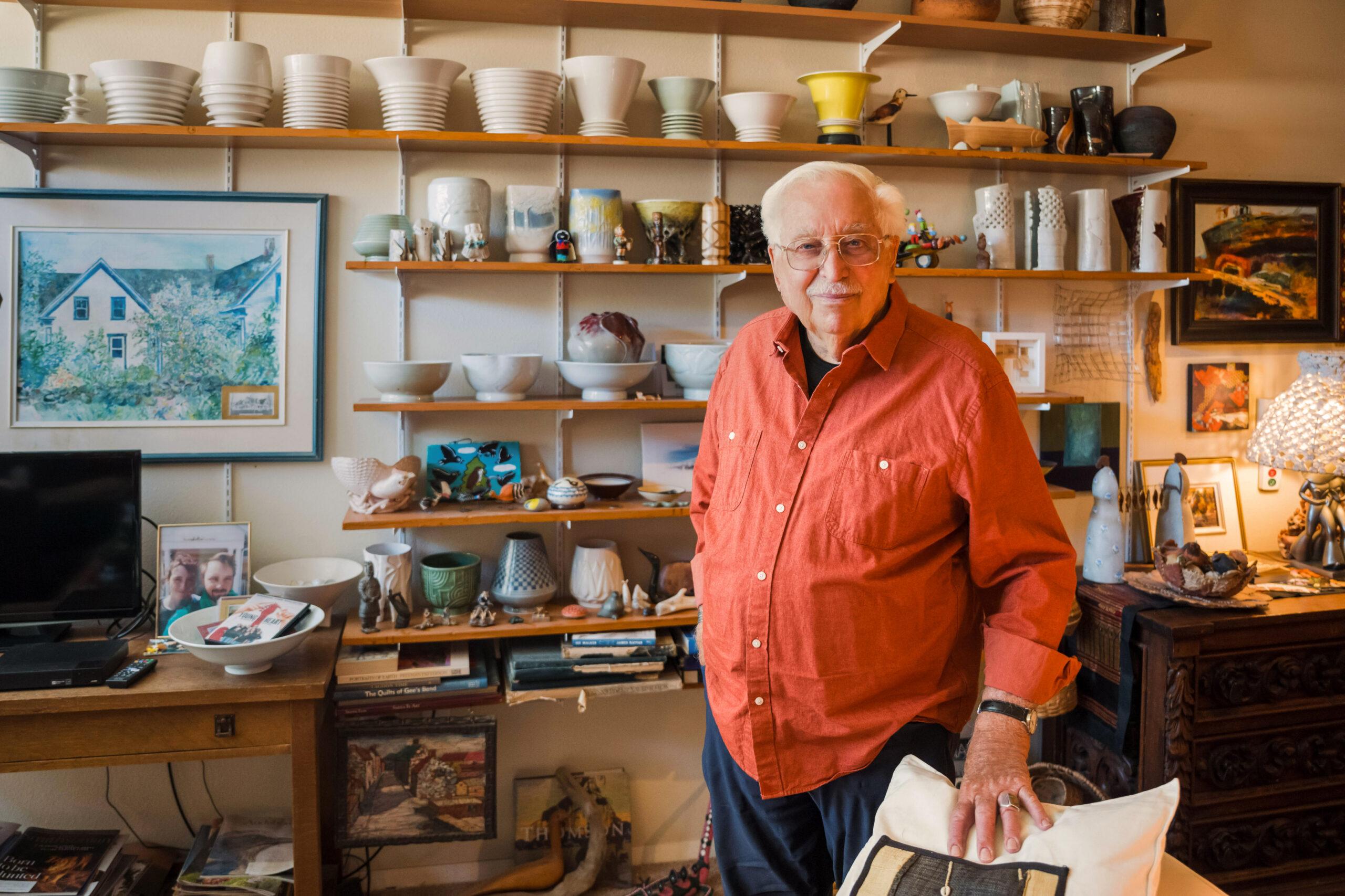 An elderly man in glasses and a red button-up shirt standing in a home studio surrounded by ceramic bowls and decorative vases.