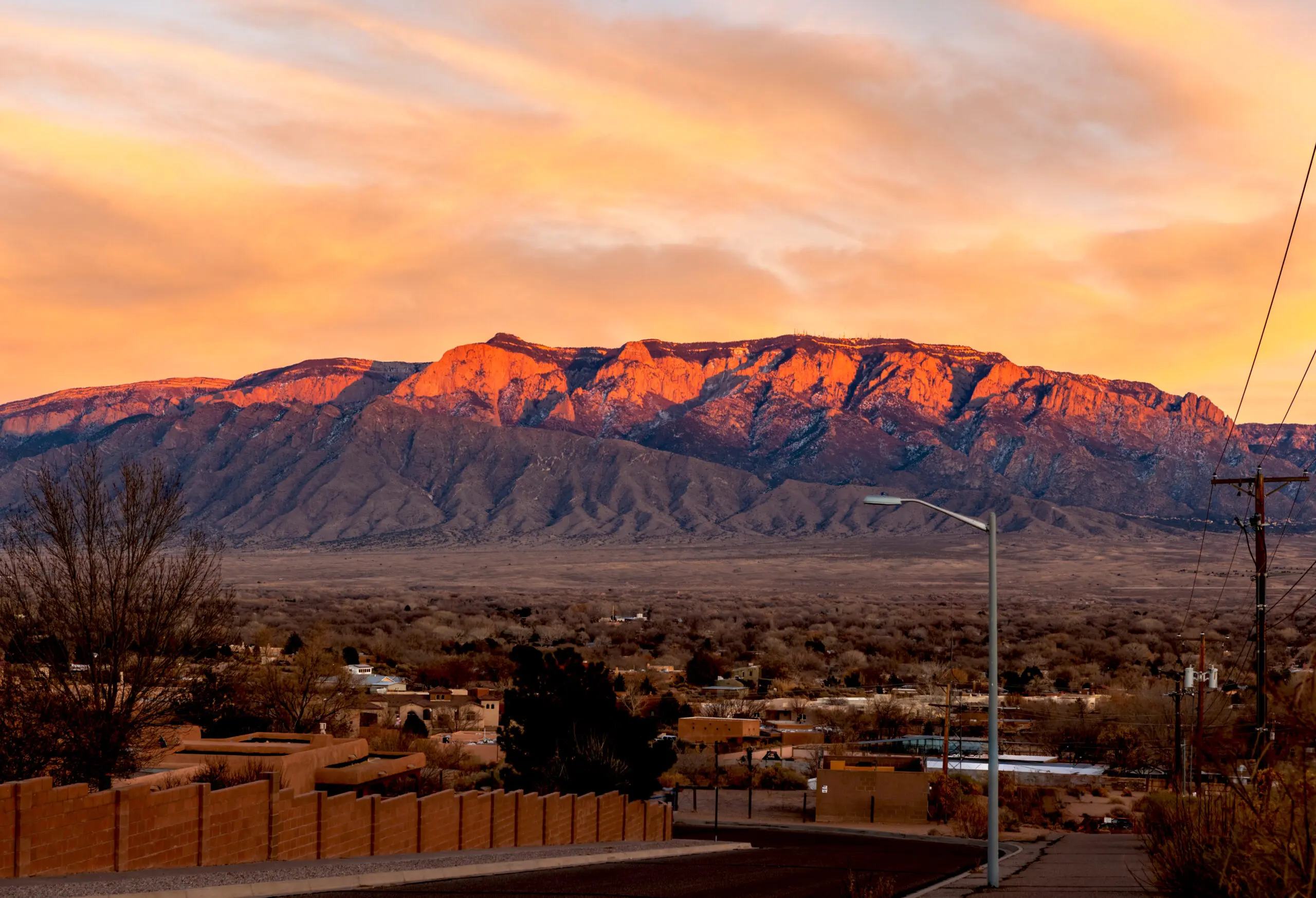 Panoramic sunset view of the Sandia Mountains glowing orange-pink against a purple sky above a sparsely vegetated desert plain.