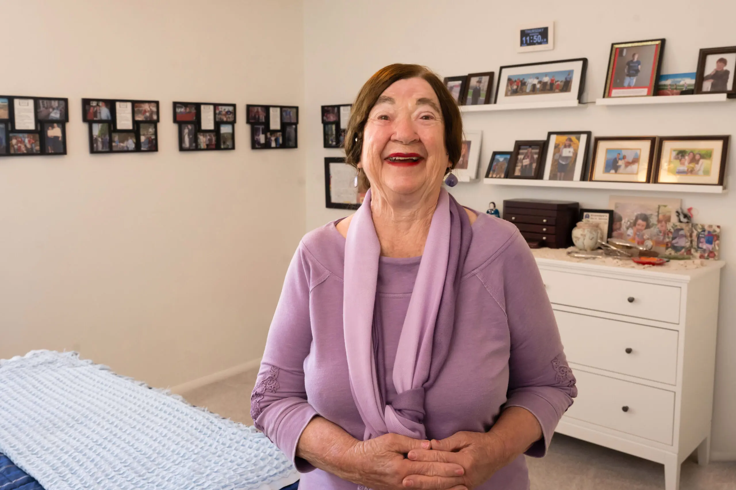 An older woman wearing a lavender top standing in a brightly lit room with family photos on shelves smiling at the camera.