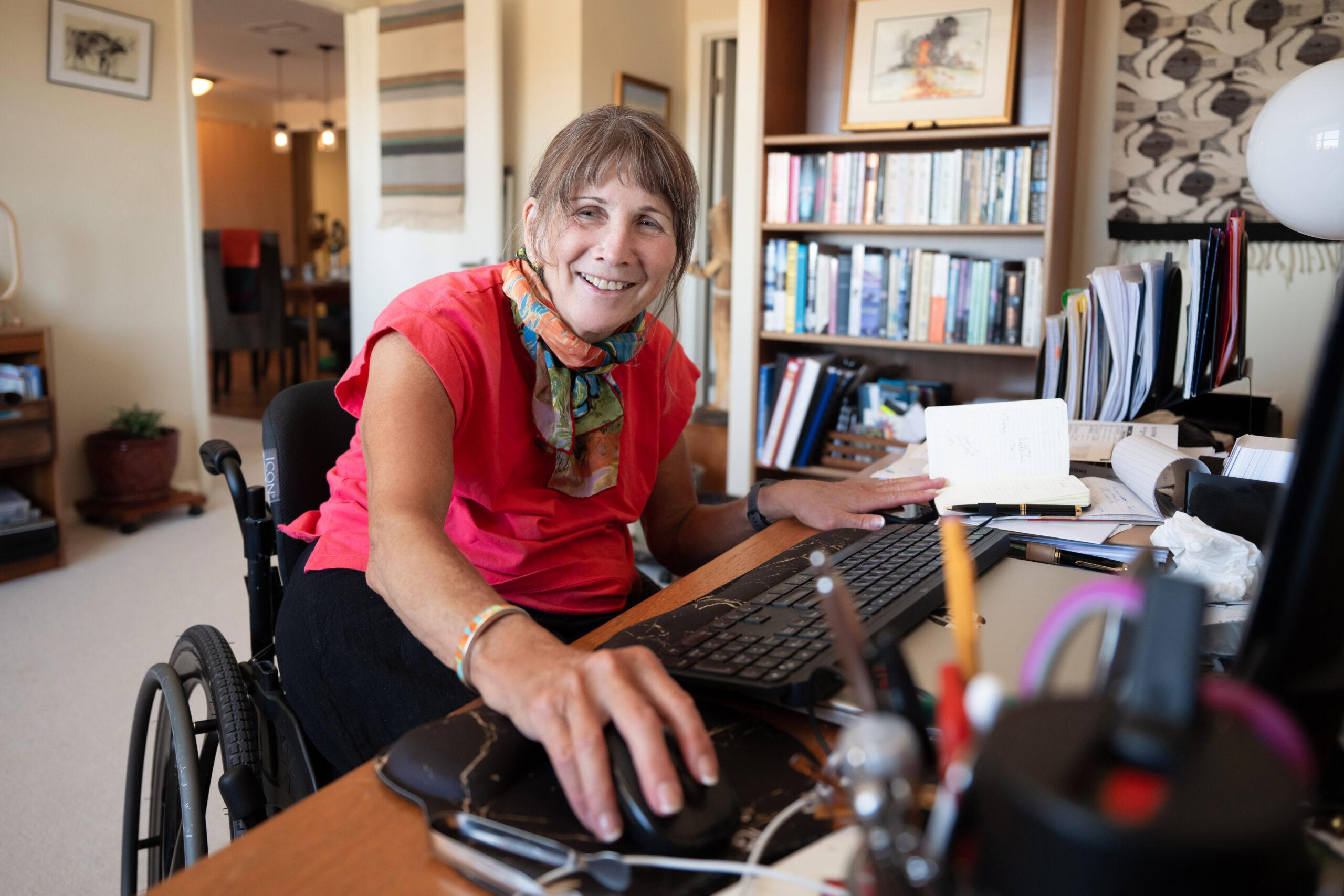 A woman seated in a wheelchair at a desk, using a computer mouse and smiling in a home office setting.