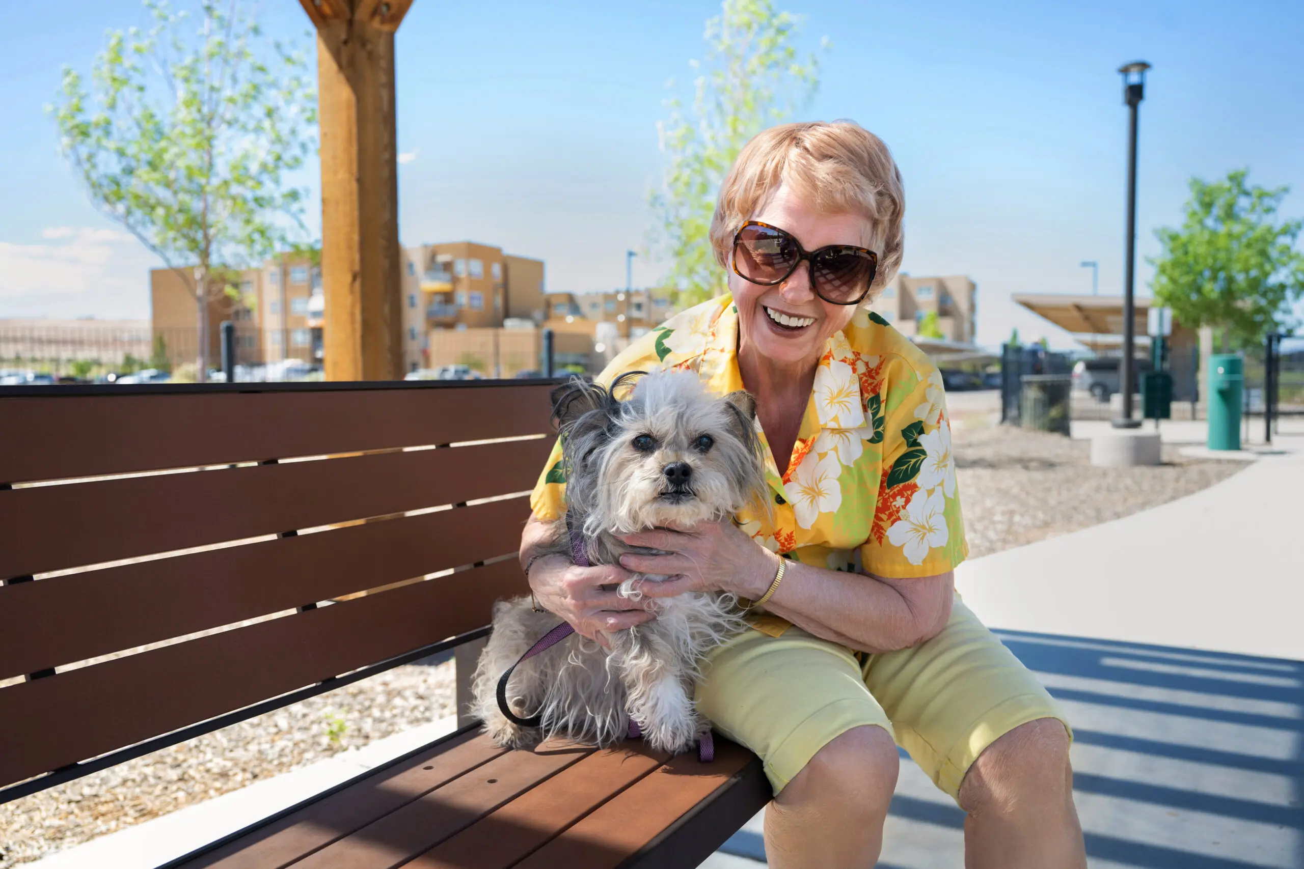 A woman sitting on a wooden bench outside holding a small fluffy dog on her lap and smiling at the camera.