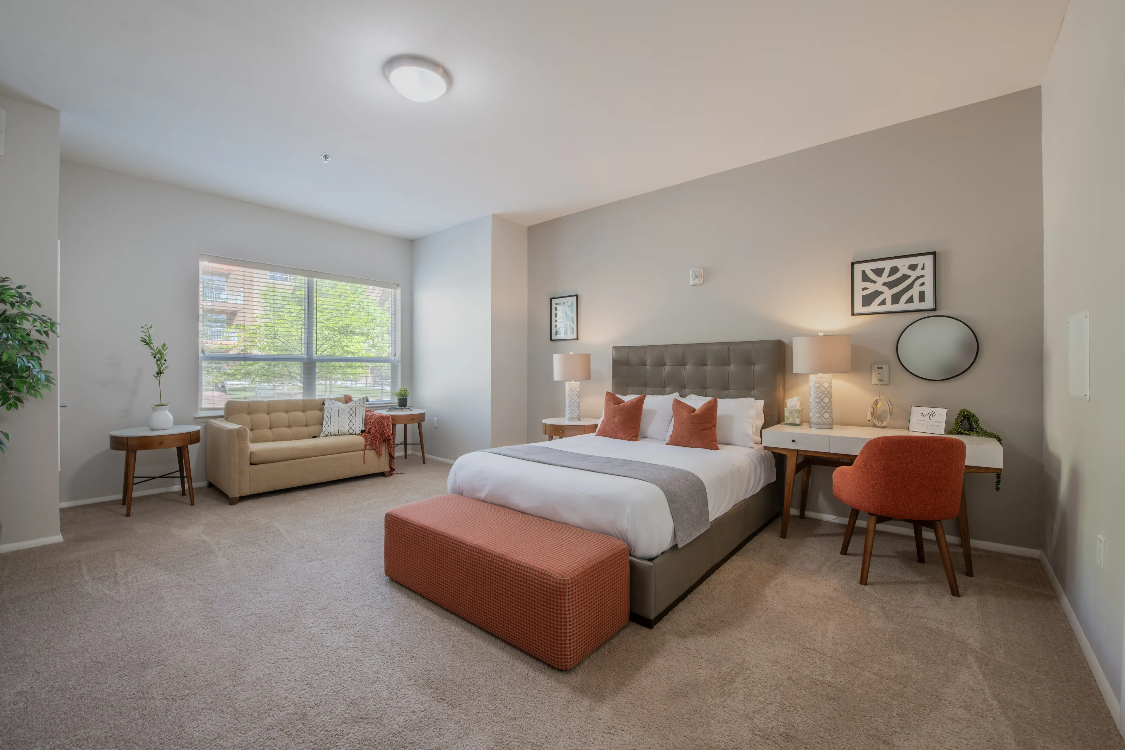 Bright apartment bedroom with upholstered bed, sofa, desk, and large window.