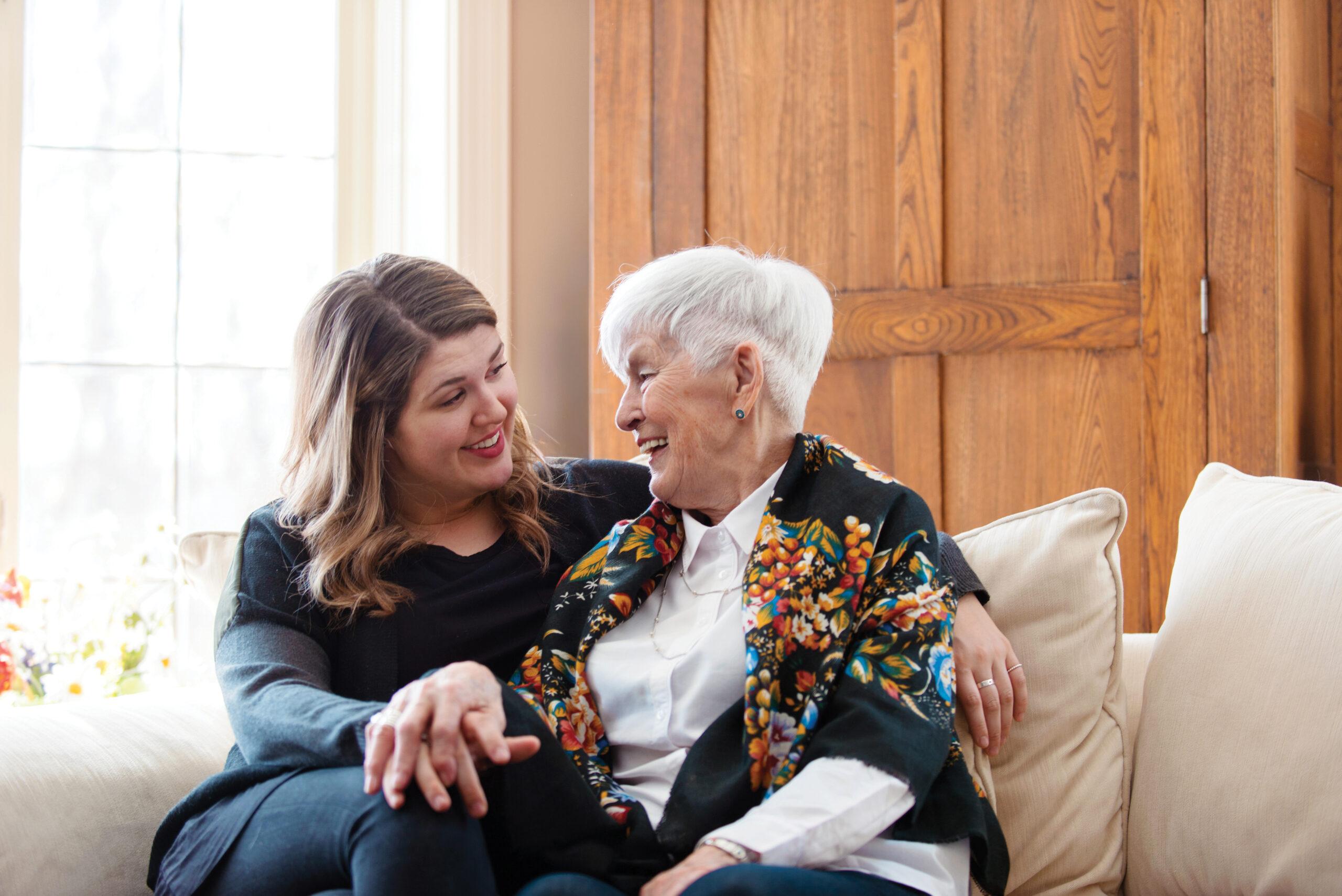 Younger caregiver and older woman sit close on a sofa, smiling and holding hands.