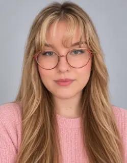 Headshot of a woman with blonde hair, bangs, and rose gold glasses, wearing a pink sweater.